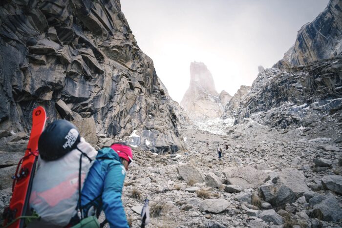 Astorga with skis on her backpack, walking up a rock debris gully towards the trango Tower, rising in background. 